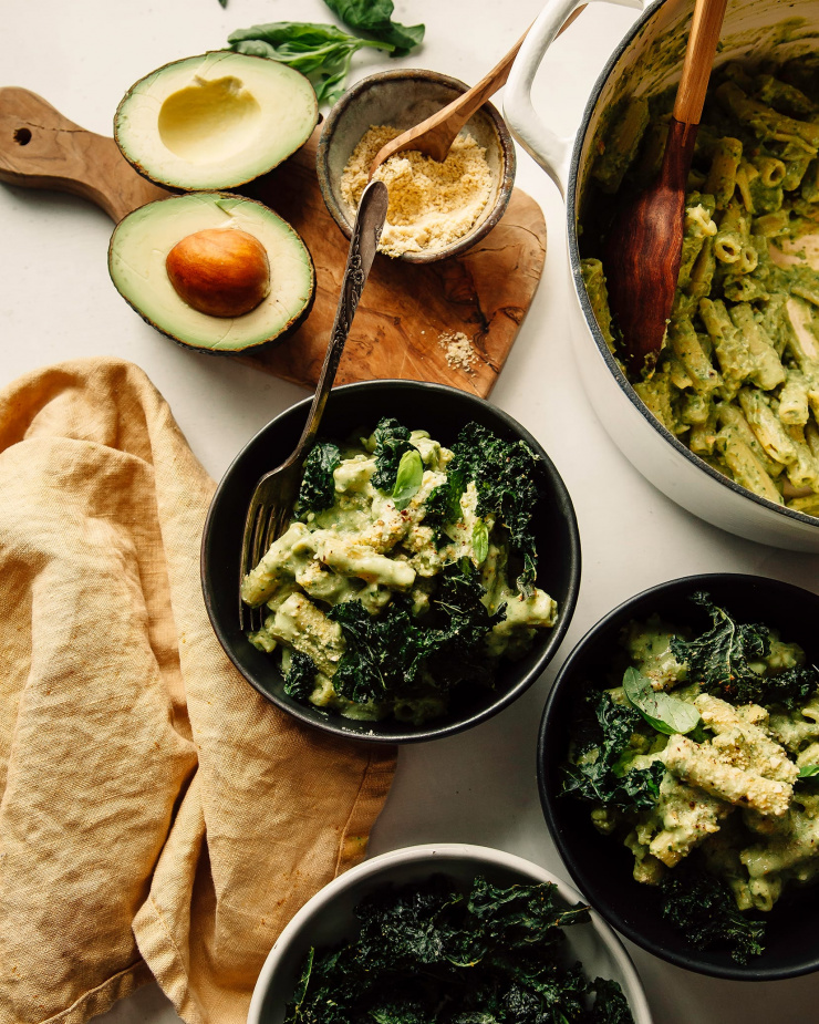 An overhead shot of avocado pesto pasta in a dark green bowls with a yellow linen nearby. The pasta is garnished with crispy pieces of kale.