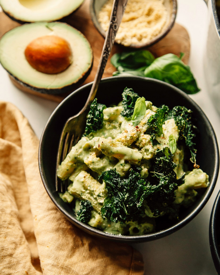 An overhead shot of avocado pesto pasta in a dark green bowl with a yellow linen nearby. The pasta is garnished with crispy pieces of kale.