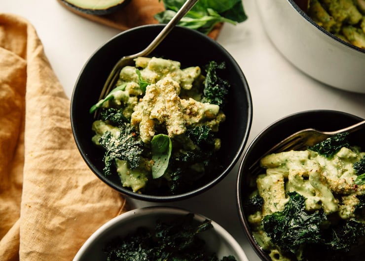 An overhead shot of avocado pesto pasta in a dark green bowl with a yellow linen nearby. The pasta is garnished with crispy pieces of kale.
