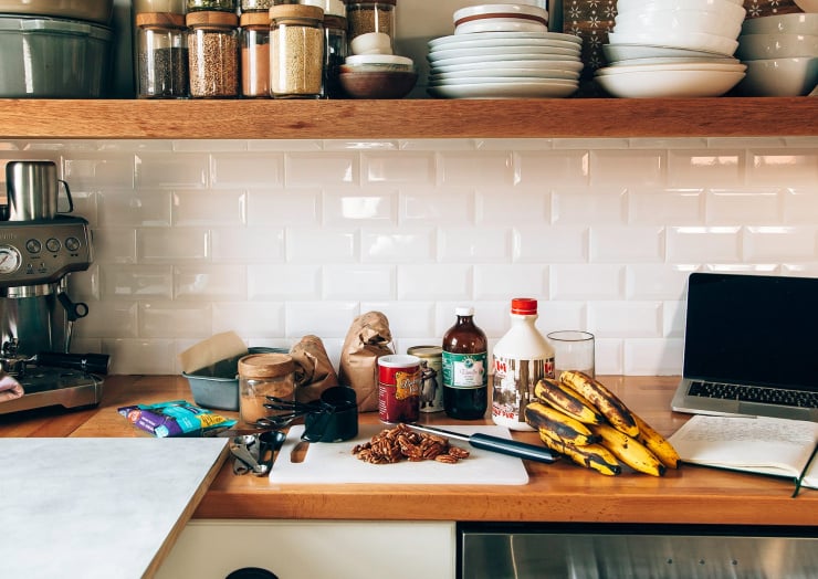 A head-on shot shows a kitchen counter with ingredients needed for making banana bread. There is also a coffee machine and laptop on the counter.