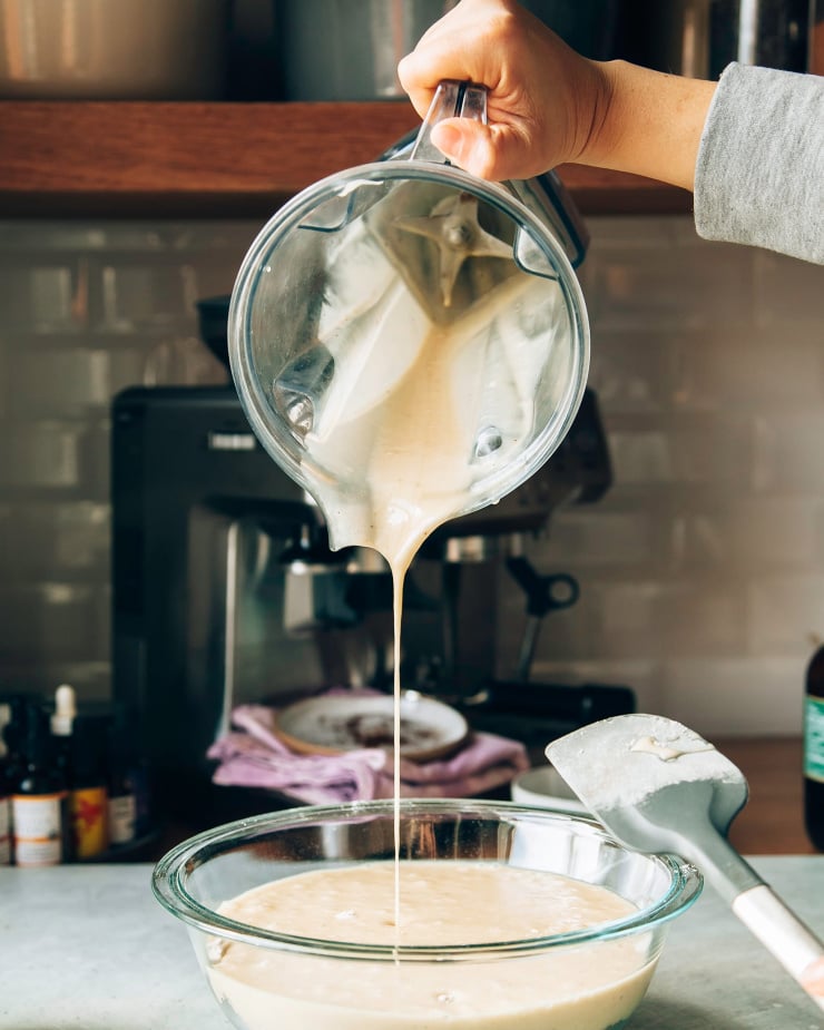 A head-on shot shows a hand pouring a blender pitcher filled with a puréed banana mixture into a mixing bowl.