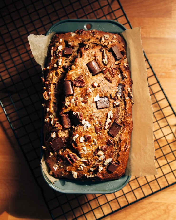 An overhead shot shows a loaf of vegan banana bread in a loaf tin, cooling on a wire rack. The top is studded with pecans and chocolate chunks.