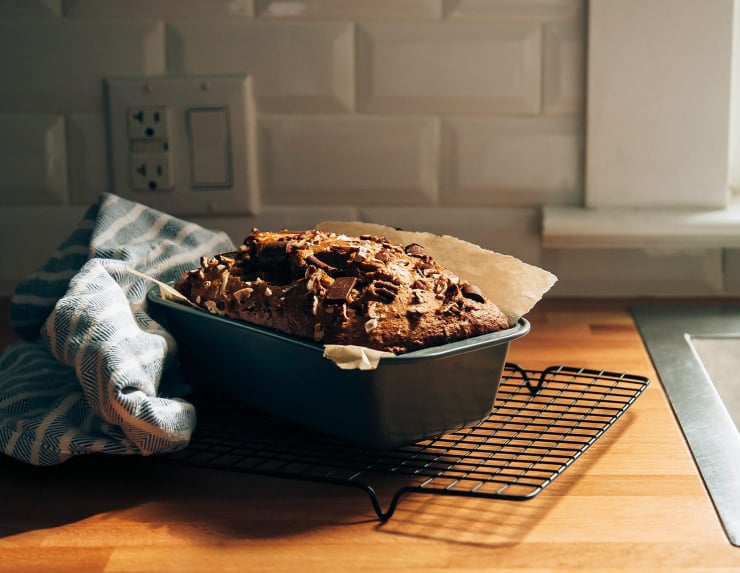A head-on shot shows a loaf of vegan spelt banana bread still in the loaf pan cooling on a wire rack.
