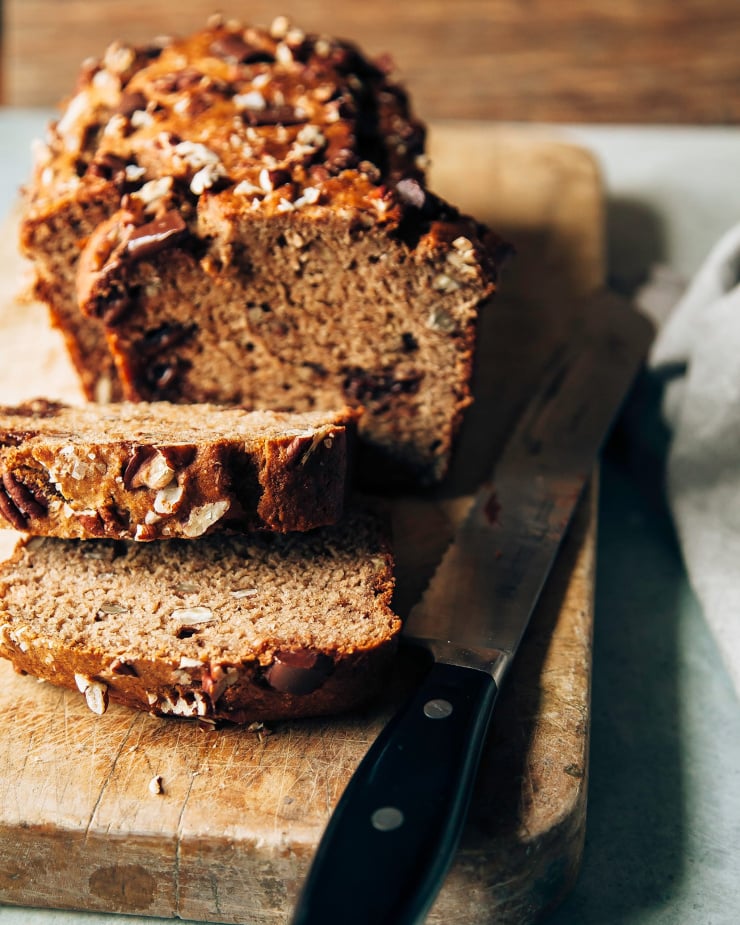 A head-on shot shows a sliced vegan spelt banana bread on a wood cutting board. A serrated knife is shown nearby.