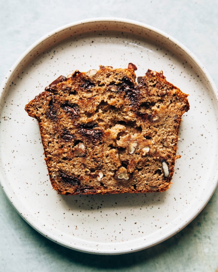 An overhead shot shows a perfect slice of vegan banana bread on a plate. You can see melted pools of chocolate and chopped pecans in the slice.