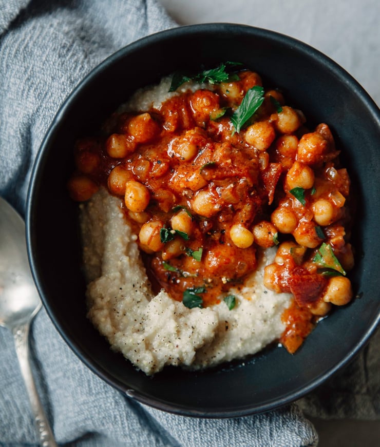 An up close, overhead shot of a deep red seven spice chickpea stew served over some creamy millet polenta. The dish is served in a matte black bowl on top of a grey linen napkin. The stew is garnished with chopped parsley.