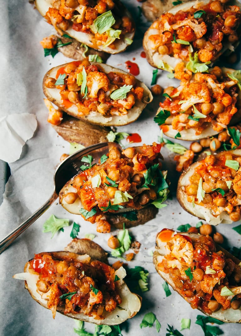 An overhead shot of baked potato halves stuffed with chickpeas and small cauliflower florets that are coated in a bright orange, Buffalo-style hot sauce. Each half is topped with chopped parsley leaves and celery leaves.