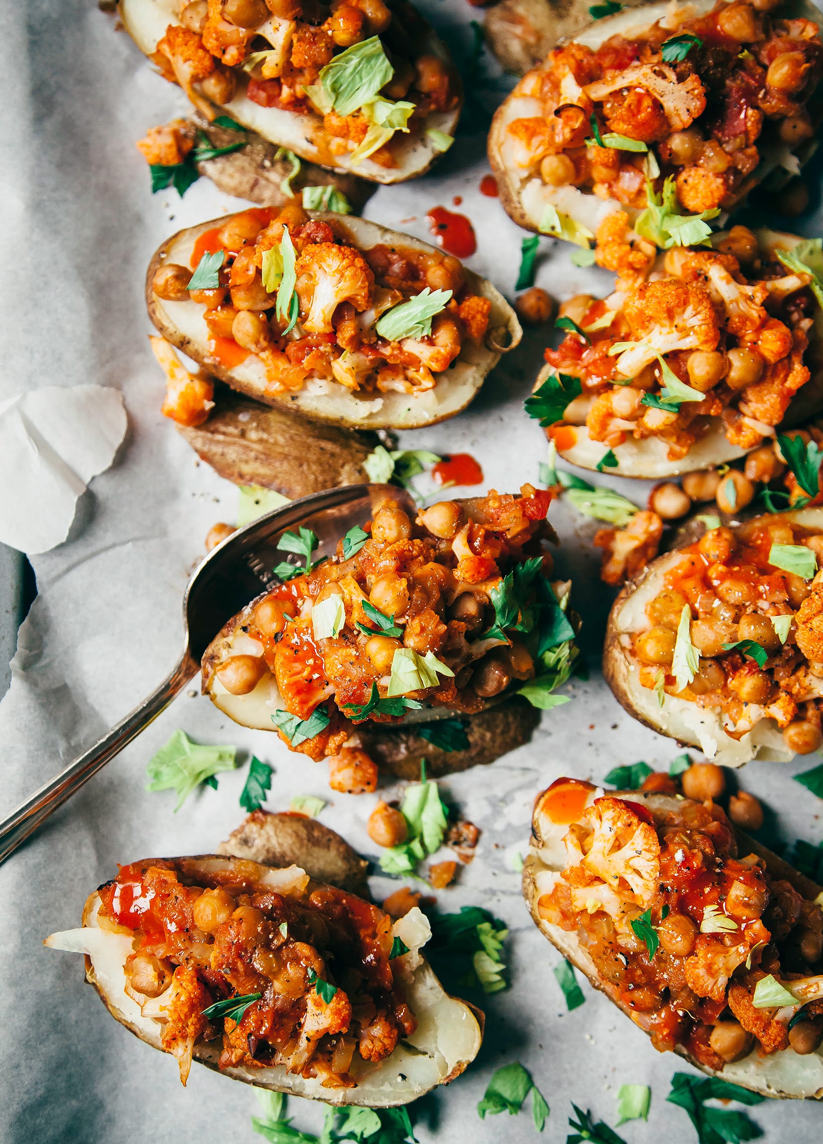 An overhead shot of baked potato halves stuffed with chickpeas and small cauliflower florets that are coated in a bright orange, Buffalo-style hot sauce. Each half is topped with chopped parsley leaves and celery leaves.