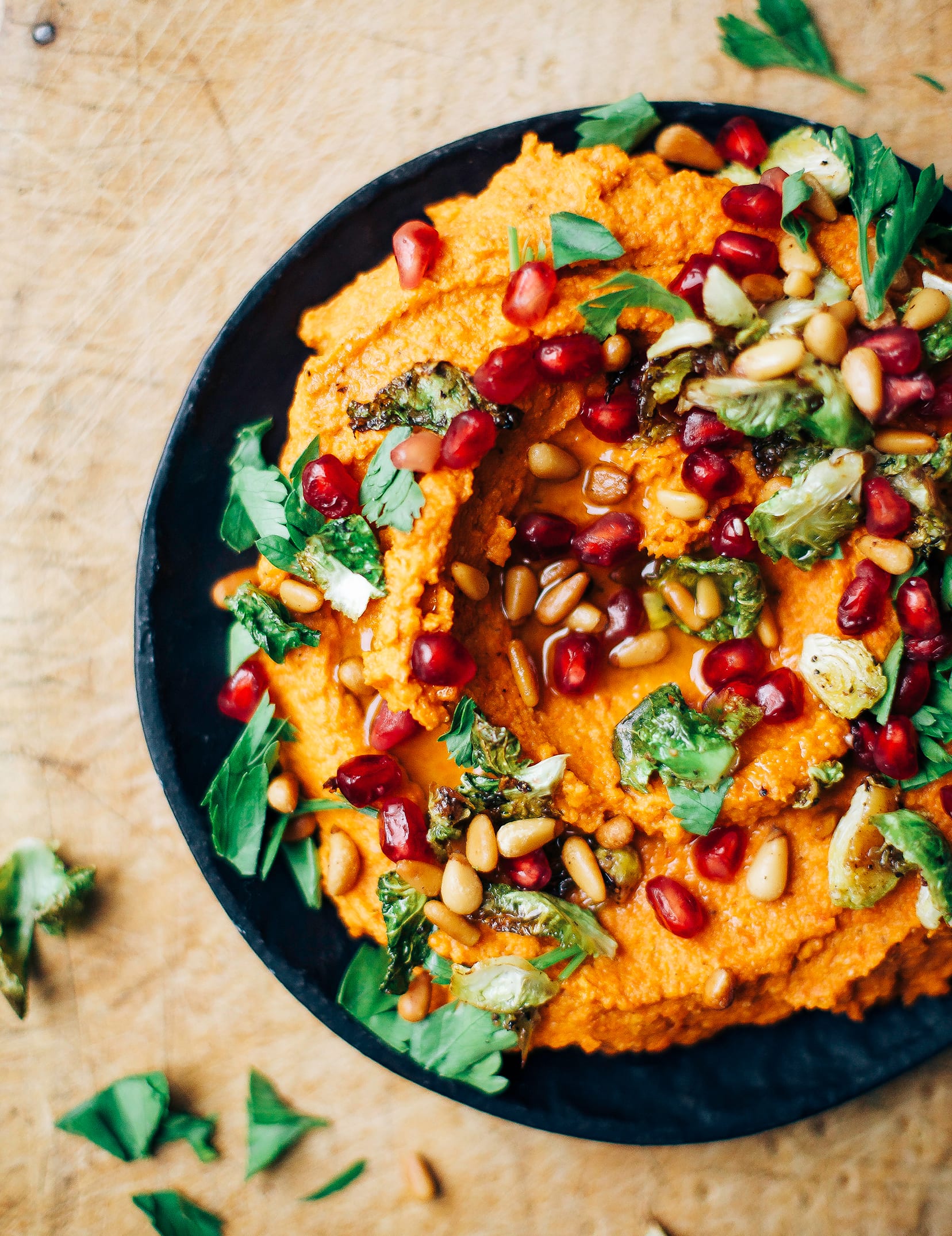 An up close, overhead shot of a bright orange dip on a black plate. The dip is topped with chopped parsley, crispy brussels sprouts, and pomegranate seeds.