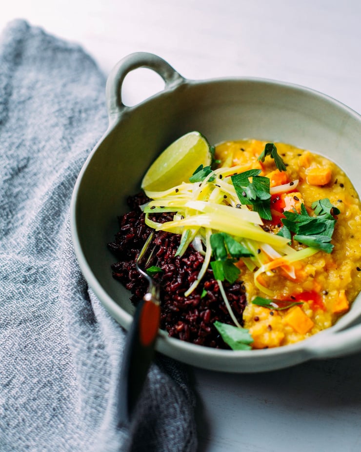 A slightly angled overhead shot of a pale orange, slightly chunky soup served with cooked leaks on top and a side of black rice. The meal is in a sage green bowl.