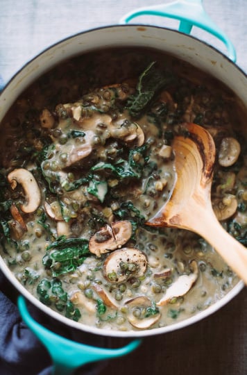 An overhead shot of a beige stew in a turquoise pot. The stew contains sliced mushrooms, kale, and cooked lentils. A wooden spoon is sticking out of the pot.