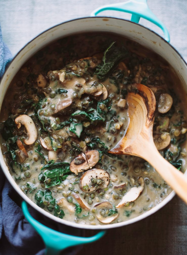 Overhead shot of creamy French lentils with mushrooms and kale in a small turquoise pot with a blue napkin. A wooden serving spoon is scooping up a bit.