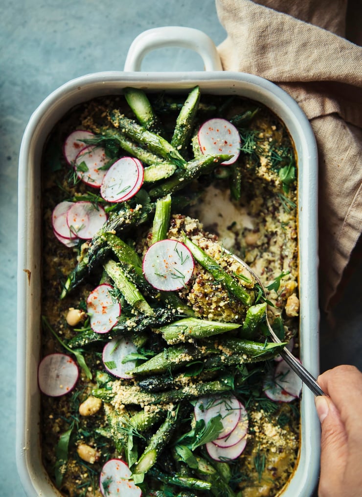 An overhead, up close shot of a hand using a spoon to serve up a portion of a quinoa and white bean casserole that is topped with sliced radishes and segments of roasted asparagus. Part of a roundup of vegan dinner recipes.