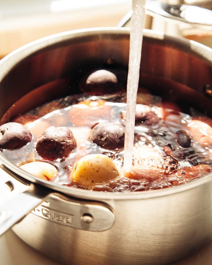 A 3/4 angle shot shows water pouring into a saucepan of baby potatoes in bright, direct sunlight.