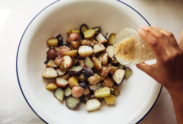 Overhead image shows a hand pouring vinaigrette from a jar onto a bowl of quartered mini potatoes.