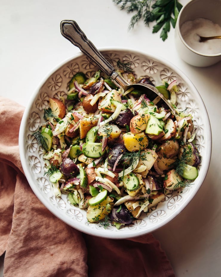 An overhead shot of a vegan potato salad with a vinaigrette dressing and herbs in a white bowl. A pink napkin is seen to the side.