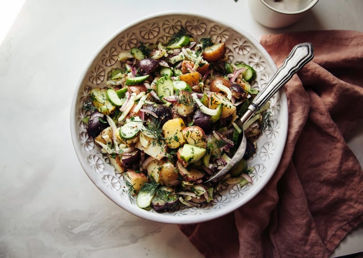 An overhead shot of a vegan potato salad with a vinaigrette dressing and herbs in a white bowl. A pink napkin is seen to the side.