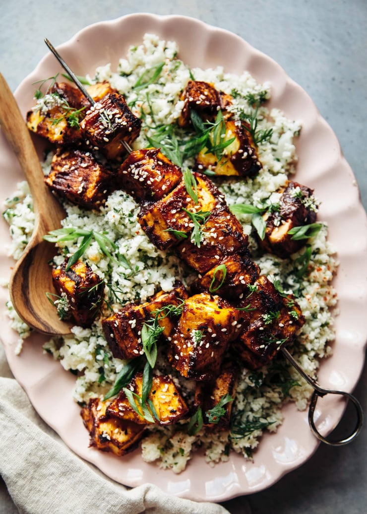 An overhead shot of a pink platter loaded up with herb-flecked cauliflower rice and skewers of grilled tofu and pineapple brushed with a caramelized, deep red sauce.
