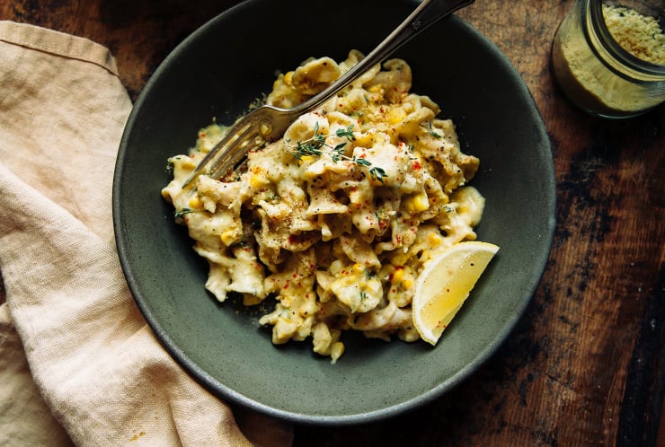 Image shows an overhead shot of a bowl of pasta on top of a worn wood background. The pasta is creamy and pale yellow, and garnished with a slice of lemon and bright red ground chili flecks.