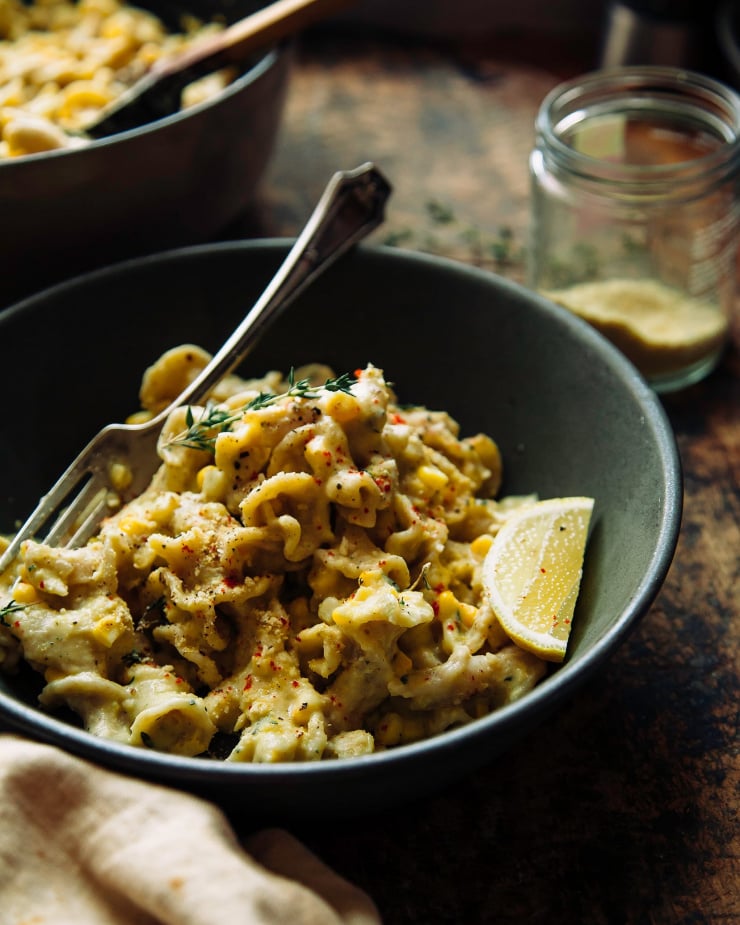 Image shows a 3/4 angle shot of a bowl of pasta on top of a worn wood background. The corn and lemon pasta is creamy and pale yellow, and garnished with a slice of lemon and bright red ground chili flecks.