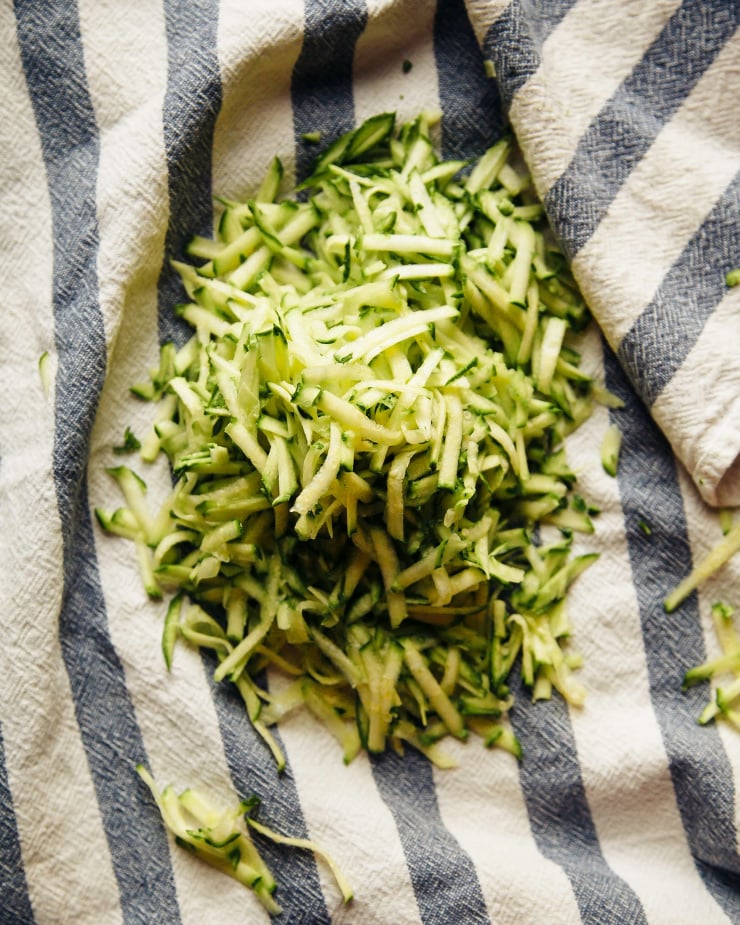 An overhead shot shows a pile of shredded zucchini in the middle of a linen towel.