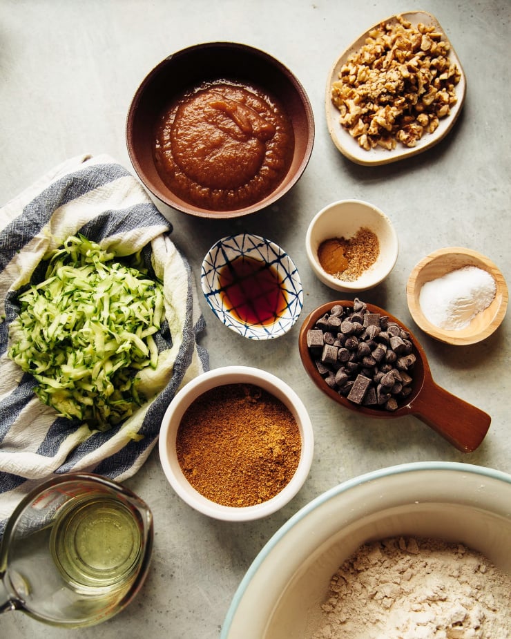 An overhead shot shows ingredients for vegan zucchini bread: apple sauce, walnuts, spices, vanilla, chocolate chips, baking powder, baking soda, coconut palm sugar, flour, oil, and shredded zucchini.