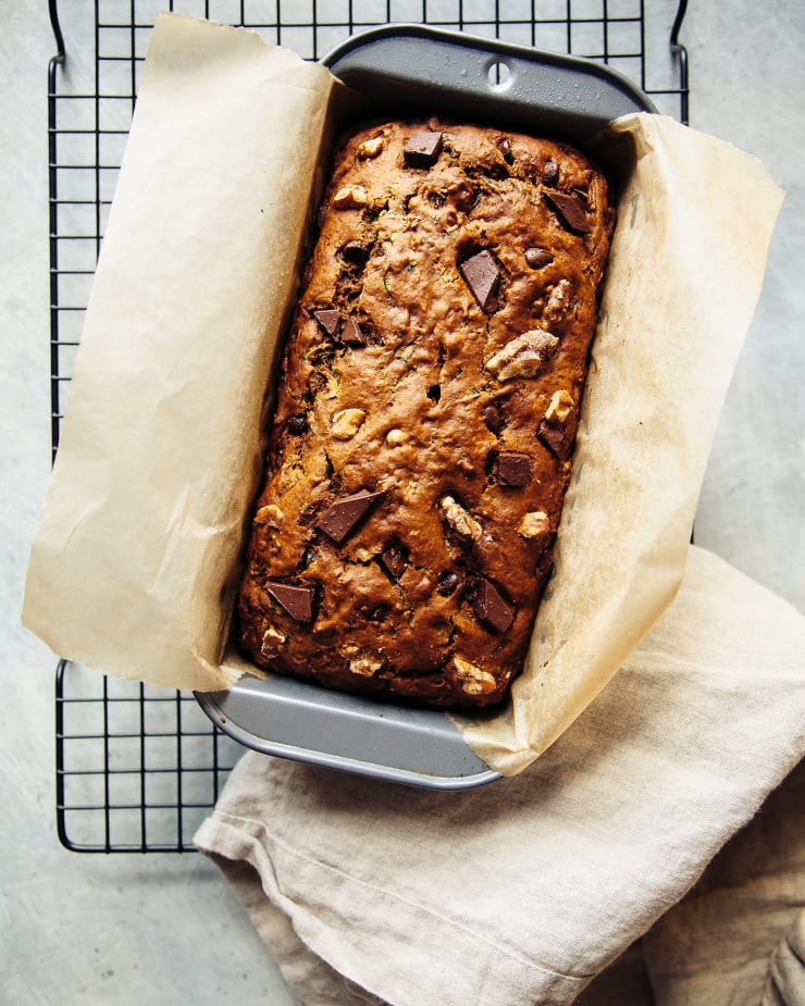 An overhead shot shows a baked vegan zucchini bread in a loaf pan on a cooling rack.