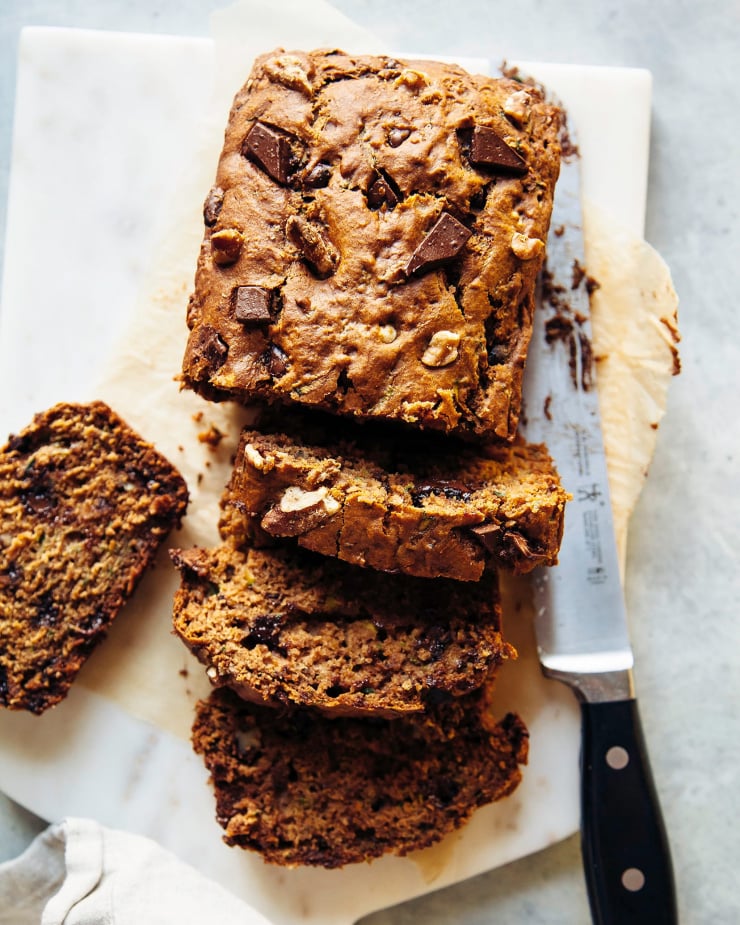 An overhead shot shows a sliced vegan zucchini bread on a white stone cutting board. The loaf is studded with chocolate and walnuts.