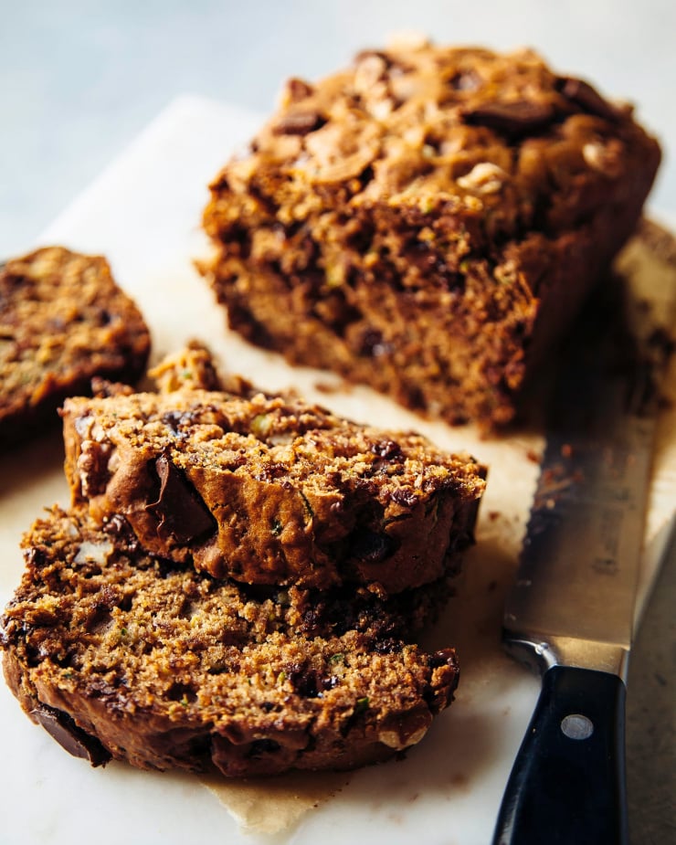 A 3/4 angle shot shows a sliced loaf of zucchini bread with a serrated knife nearby. Gooey spots of chocolate can be seen in the slices.