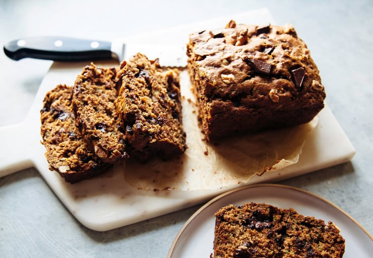 A 3/4 angle shot shows a sliced loaf of zucchini bread with a serrated knife nearby. Gooey spots of chocolate can be seen in the slices.