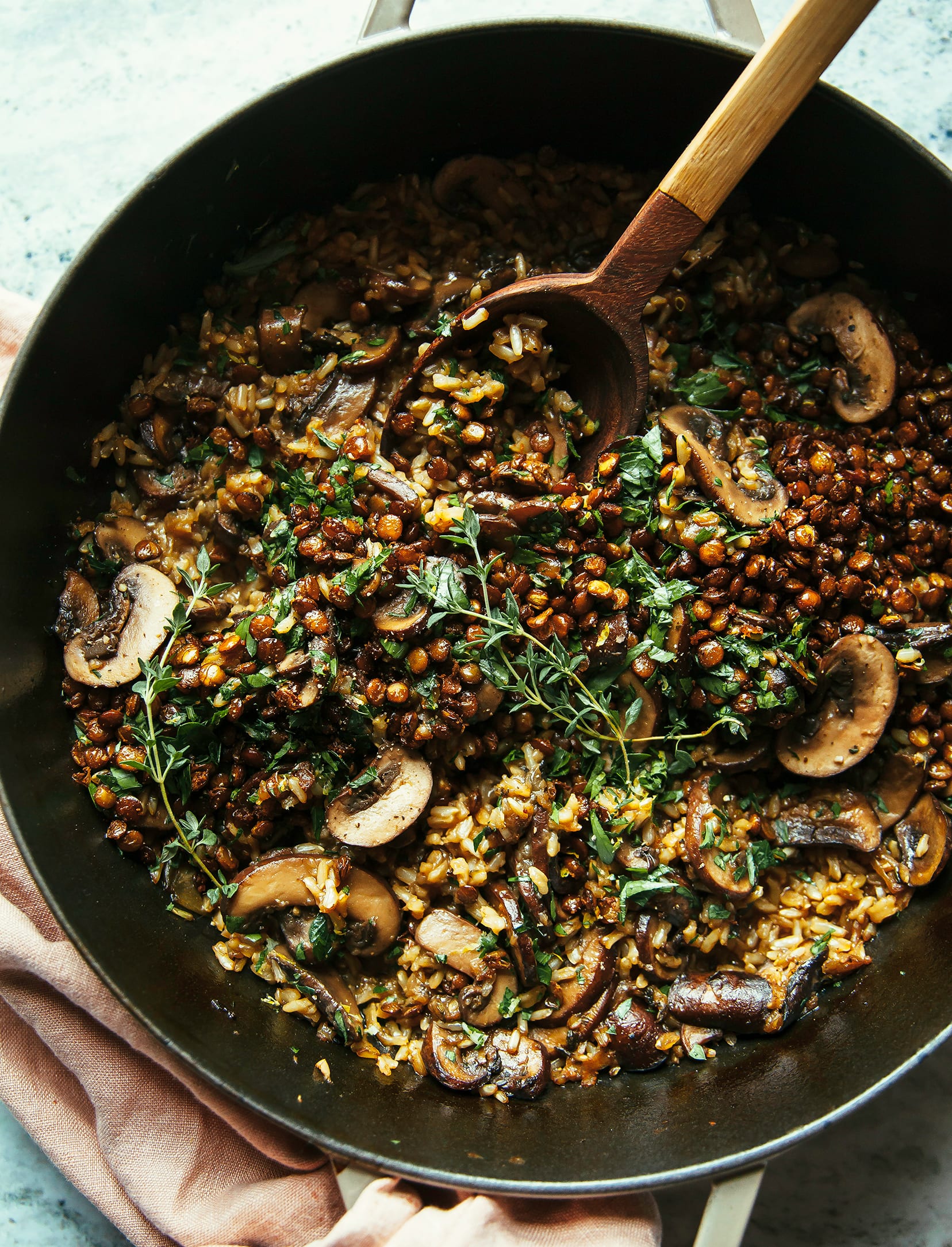 An overhead shot of a brown rice bake with lentils, shallots, and herbs. The bake is photographed in a braiser-style pot with a wooden spoon sticking out of the pot.