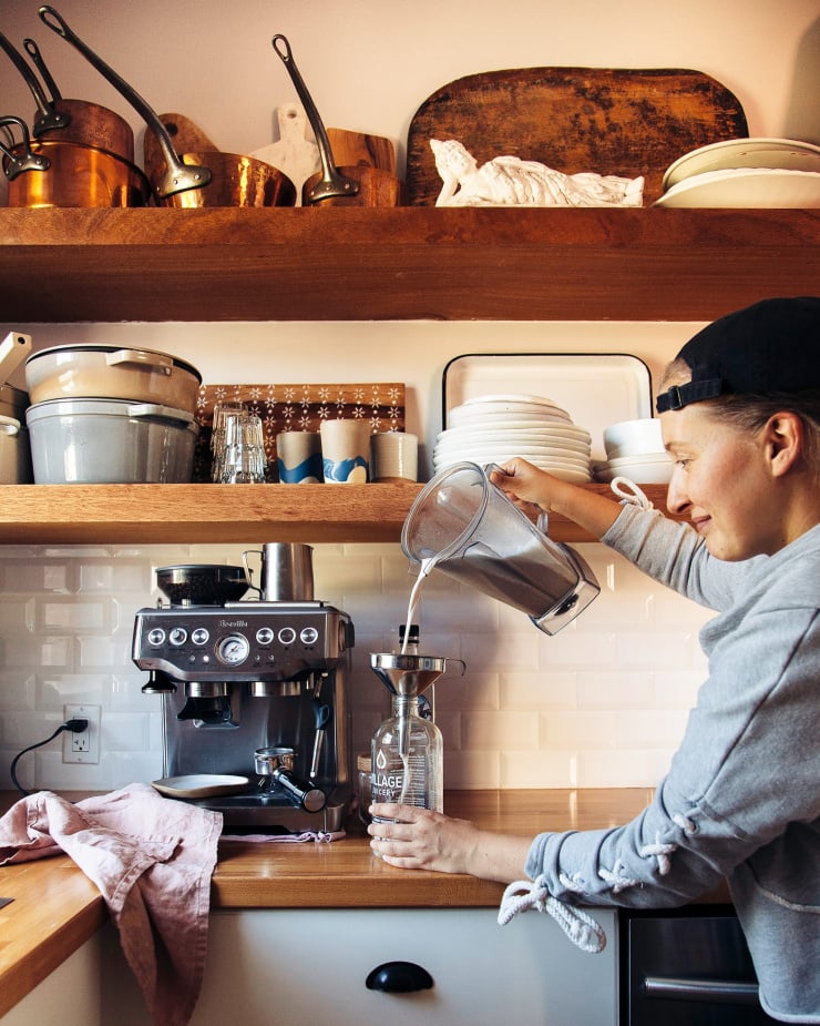 A woman is seen pouring a creamy substance from a blender pitcher into a glass bottle. A kitchen with open shelving and wood counters is seen in the background.