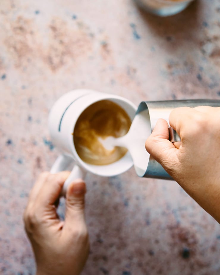 A hand is pouring frothed milk into a cup of coffee.