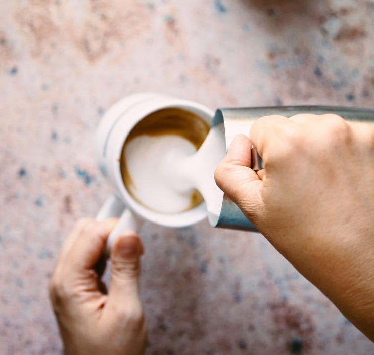 A hand is pouring frothed milk into a cup of coffee.