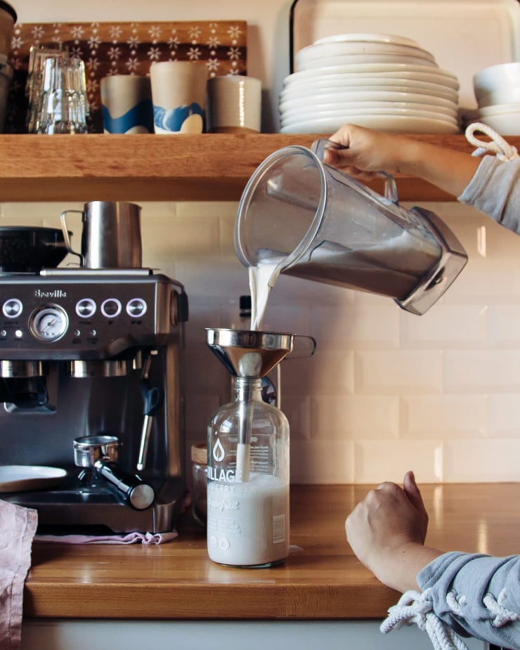 A hand is pouring hemp milk from a blender pitcher into a bottle. A kitchen is seen int he background with open shelving and a coffee machine.