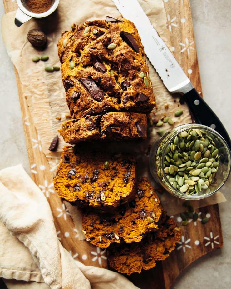 An overhead shot shows a vegan pumpkin bread loaf on a paper-lined wooden cutting board. A serrated knife is nearby and a few slices of the loaf have been cut, revealing loads of chocolate chips.