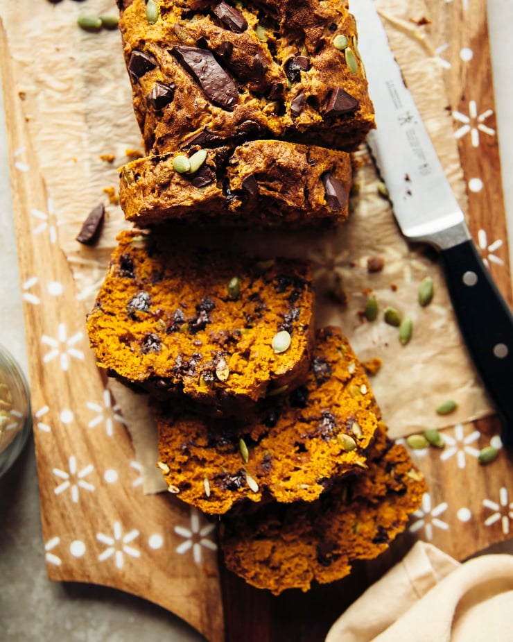 An overhead shot shows a sliced vegan pumpkin bread loaf with chocolate chips and pumpkin seeds. A serrated knife is on the board nearby.