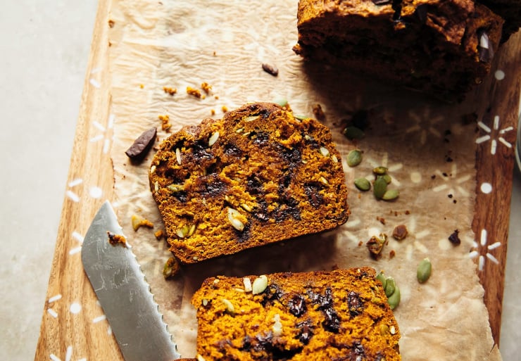 An overhead shot shows a slice of vegan pumpkin loaf studded with melty chocolate bits. It's on a cutting board lined with parchment paper with the whole loaf nearby.