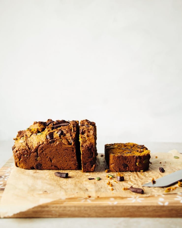 A head-on shot shows a vegan pumpkin loaf against a white background. A few slices have been cut from the loaf so far.