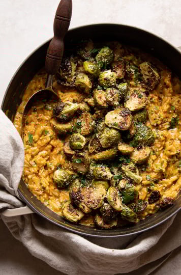 An overhead shot of a creamy vegan orzo risotto with a slight orange hue. The orzo risotto is in a braiser-style pot and topped with crispy roasted brussels sprouts.
