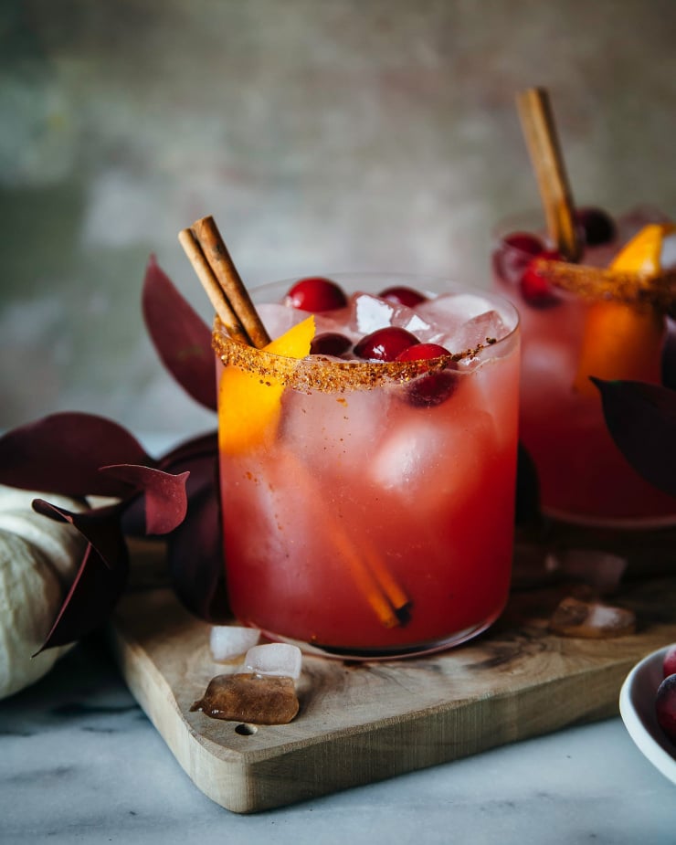 a head on shot of two cranberry orange margaritas garnished with cinnamon sticks, whole cranberries, and orange peels. The drinks are deep pink.