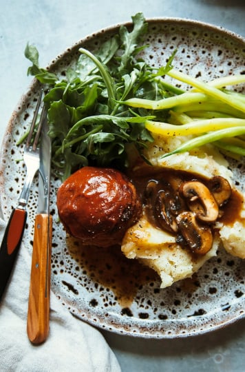 An overhead shot of a dinner plate that features one mini vegan buffalo meatloaf, some dressed arugula, a heap of mashed potatoes with mushroom gravy spooned on top, and some simply sautéed yellow string beans.