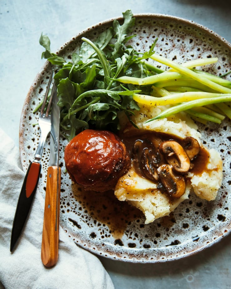 An overhead shot of a dinner plate that features one mini vegan buffalo meatloaf, some dressed arugula, a heap of mashed potatoes with mushroom gravy spooned on top, and some simply sautéed yellow string beans.