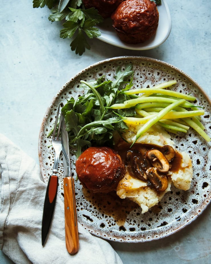 An overhead shot of a dinner plate that features one spicy vegan buffalo meatloaf mini, some dressed arugula, a heap of mashed potatoes with mushroom gravy spooned on top, and some simply sautéed yellow string beans.