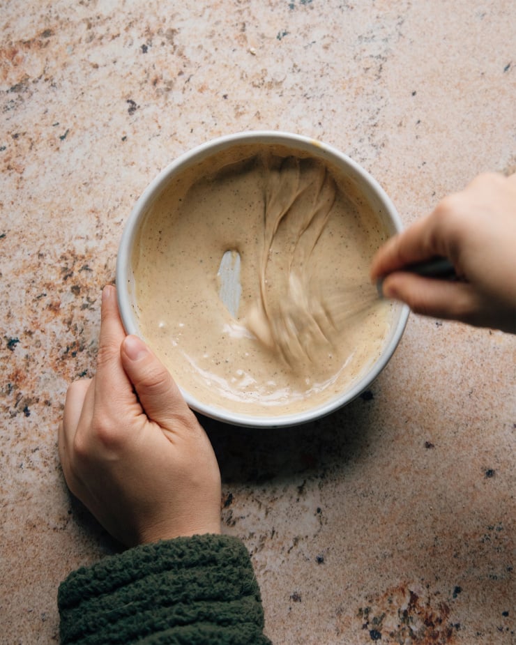 Whisking the maple tahini glaze in a white bowl on a pink, stone-like background.
