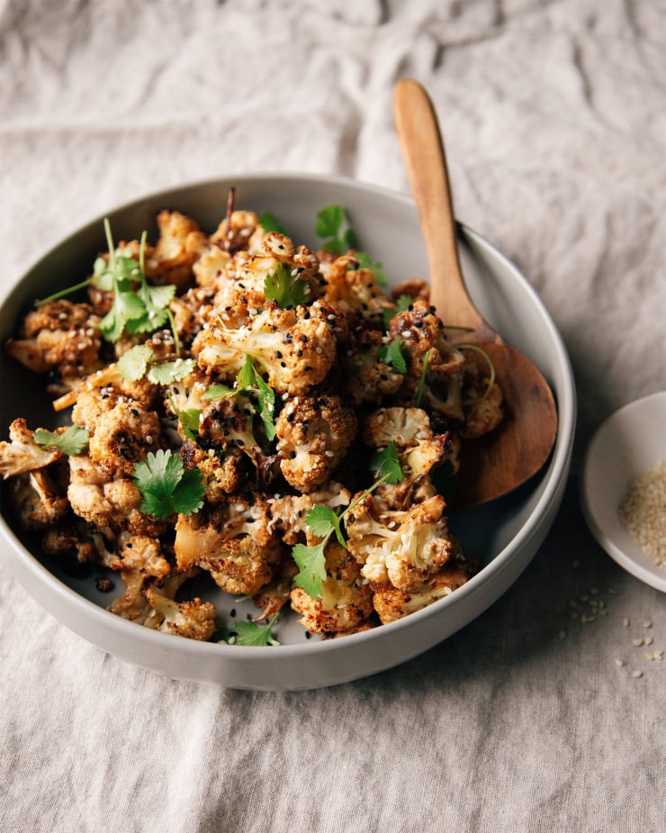 A bowl of maple tahini glazed cauliflower with chipotle. It is garnished with cilantro and sesame seeds. Shot is taken at 3/4 angle on a beige linen.
