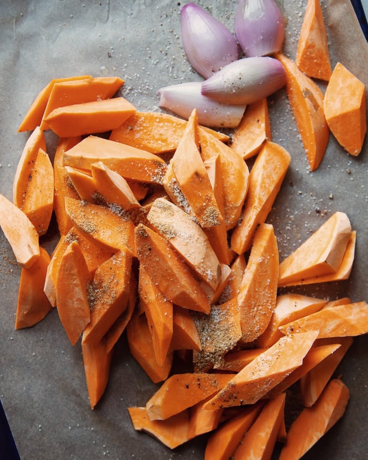 An up close shot of chopped sweet potatoes and shallots on a baking sheet with ground coriander, salt, and pepper sprinkled on top