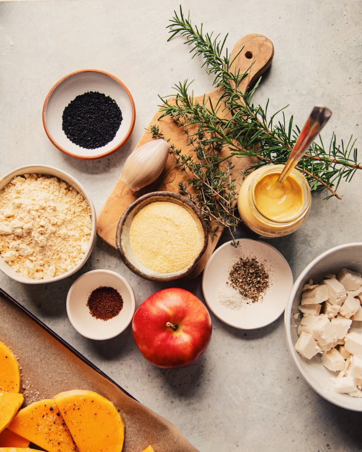 An overhead shot of ingredients, including: rosemary, thyme, squash, chickpea flour in a bowl, cornmeal in a bowl, salt, pepper, and apple, shallot, and a small bowl of nigella seeds. All on a grey background.