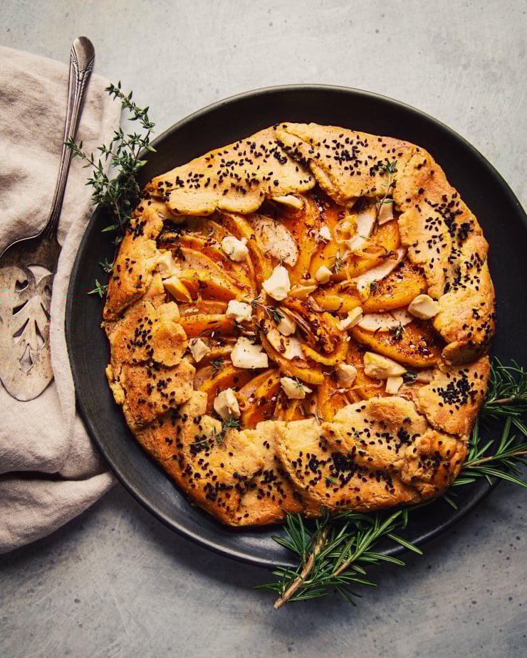 An overhead shot of a vegan butternut galette on a matte black plate on a mottled grey background. It is garnished with thyme and rosemary sprigs. There is a beige linen and antique pie server to the side.