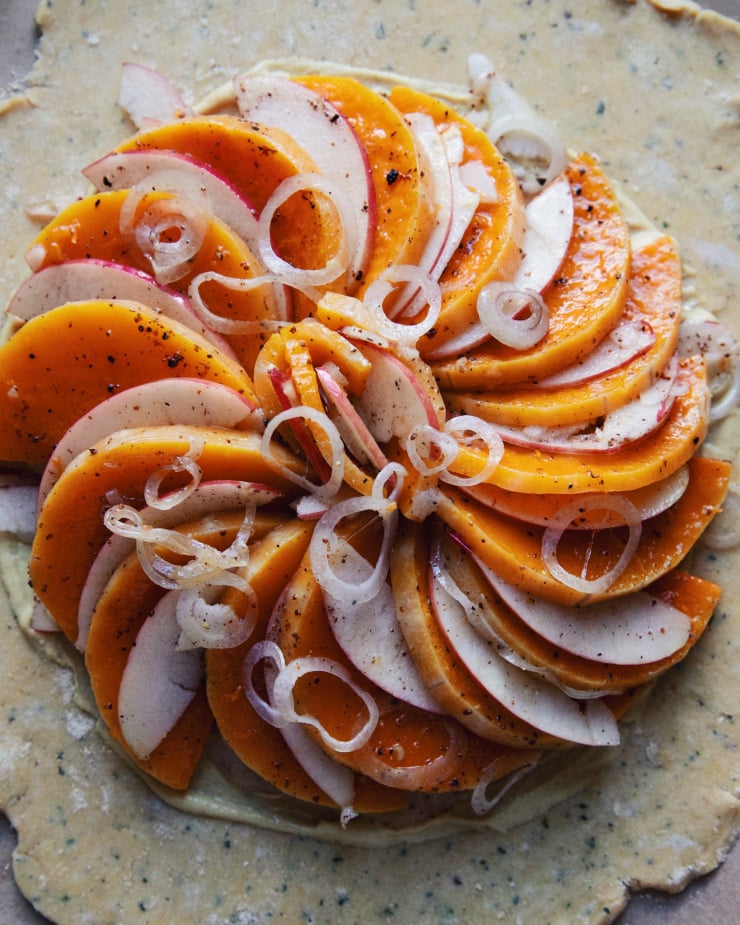 An overhead shot of a galette pre-bake. Butternut squash, apples and shallots are layered in a circular pattern over some rolled out pastry.